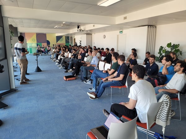 A group of people are seated in a large, modern room, attentively watching a presentation given by a speaker standing at the front.
