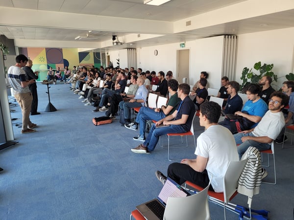 A large group of people are seated in an indoor hall, attentively listening to two men who are standing and giving a presentation.