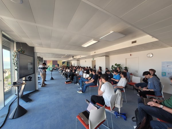 A group of people attending a presentation in a modern, bright office space with a speaker standing near a large screen.