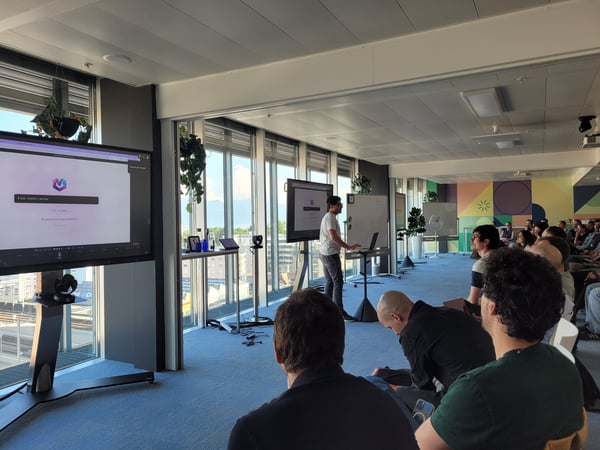 A man is giving a presentation in a modern office setting to a seated audience, with large screens displaying a software installation command.