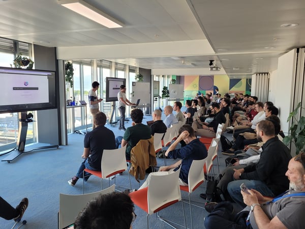 A photograph of a presentation or meeting in a modern office setting, with an audience seated in rows facing two presenters and a large display screen.