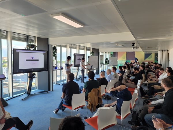 A group of people attending a technical presentation in a modern office space with large windows and multiple digital displays.