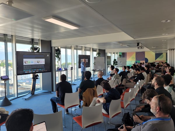 A group of people are seated in a modern office space, attending a technical presentation about the 'Segment Anything Model'. A speaker stands at the front near a large screen displaying diagrams and text, with large windows showing a city view in the background.
