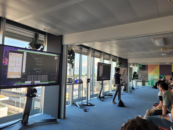 A man is giving a technical presentation in a modern office space with large windows, showing code and a mobile app interface on a large screen to an audience.