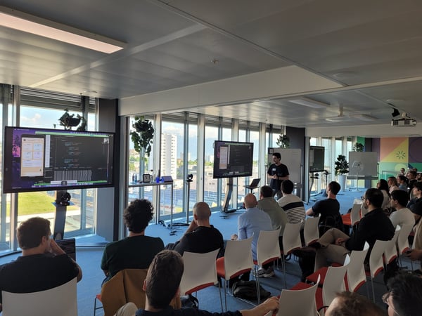 A man stands at the front of a modern office space, giving a technical presentation to an audience. Large screens display code and a mobile application interface against a backdrop of city views through large windows.