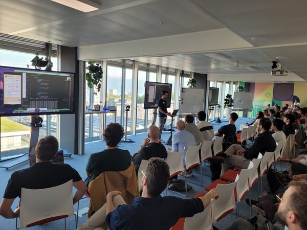 A man stands at the front of a modern office space, giving a technical presentation or software demo to a seated audience.