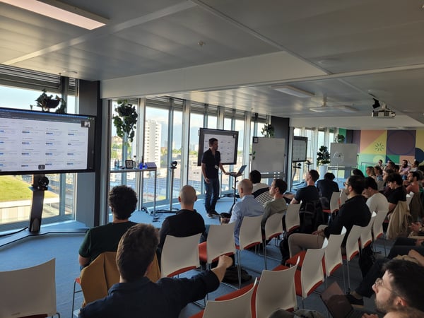 A man stands in front of a seated audience, giving a presentation in a modern office space with large windows and multiple digital screens.