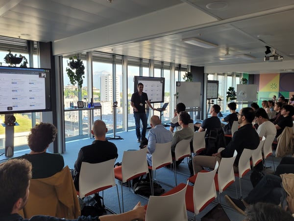 A man is giving a presentation to a seated audience in a bright, modern office space with large floor-to-ceiling windows showing a city view.