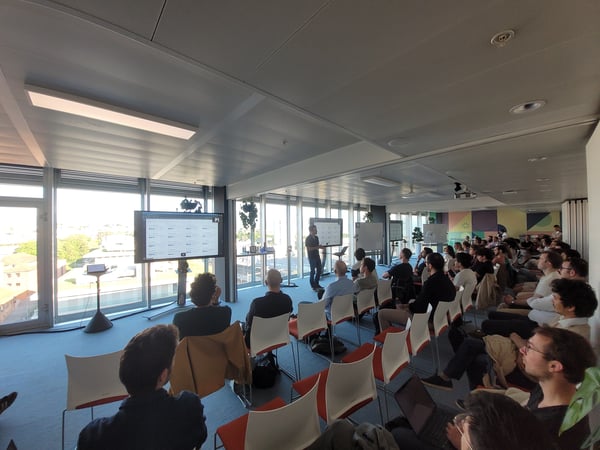 A speaker stands at the front of a modern office space, delivering a presentation to a seated audience in front of large windows.