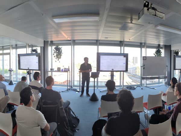 A wide-angle panoramic photo of a man giving a presentation to a large audience in a modern, bright office space with multiple large screens.