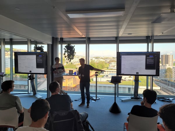 A man is giving a presentation in a modern office or conference room with large windows overlooking a city. Two large screens display a software interface while an audience sits in the foreground.