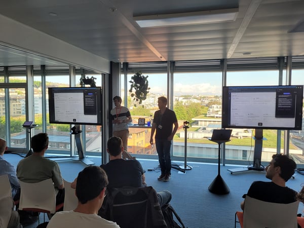 A presentation or product demo taking place in a modern office with large windows overlooking a city. Two presenters stand near large screens while an audience watches from their seats.