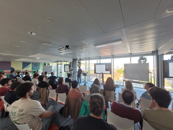 A group of people sitting in a modern office space, attending a presentation given by two individuals at the front of the room.