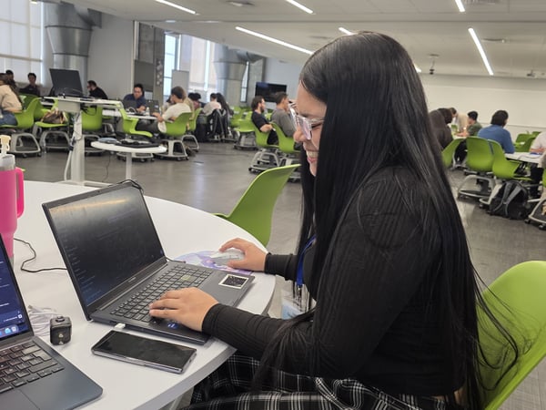 A woman with long black hair is working on a laptop in a large, open-plan workspace with several other people at tables with green chairs.