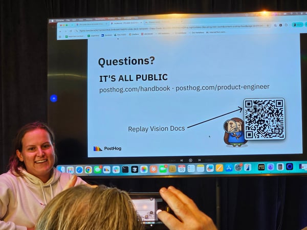 A woman stands next to a large display screen showing a PostHog presentation slide with a QR code, while an audience member in the foreground takes a photo of the screen.