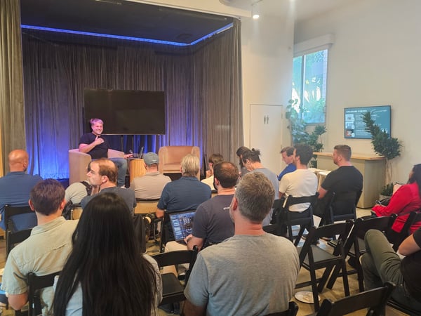 An indoor scene showing a speaker sitting on a small stage presenting to an audience seated in rows of chairs.