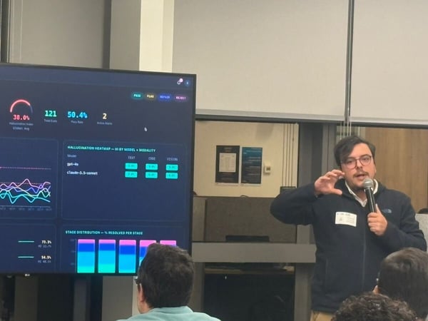A man stands presenting in front of a large monitor displaying a data dashboard, holding a microphone in one hand and gesturing with the other.