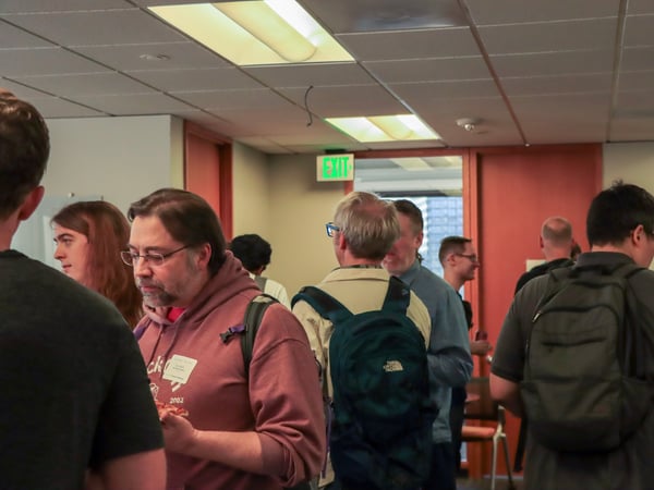A group of people are gathered in an indoor office or event space, with some individuals wearing backpacks and others engaged in conversation.