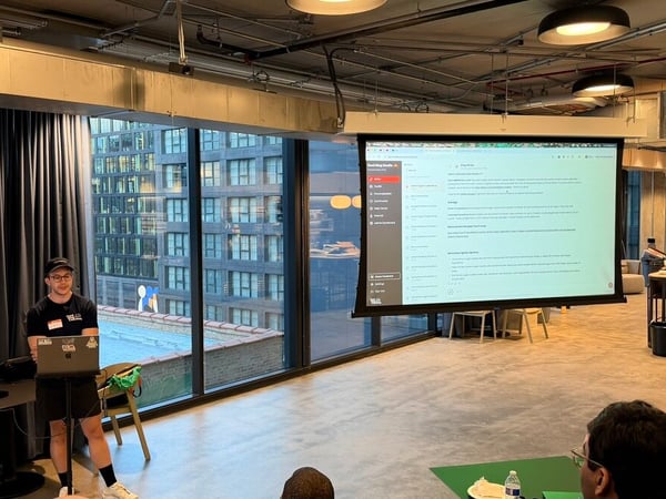 A presenter stands next to a laptop in a modern office, delivering a presentation shown on a large projector screen.