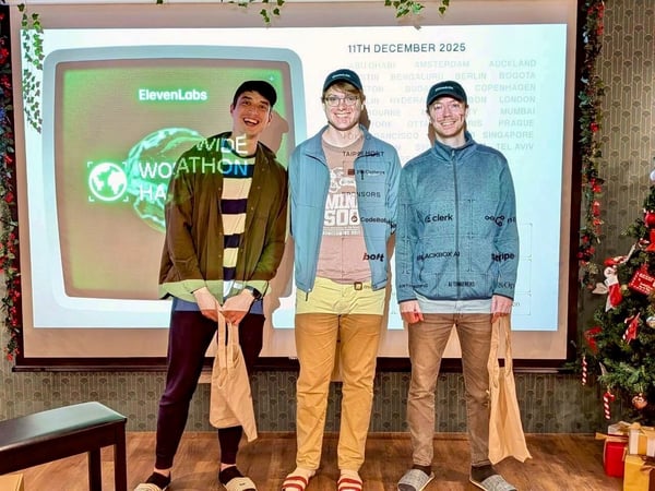A photograph showing three young men standing and smiling in an indoor setting, posing in front of a large projection screen displaying event information, logos, and a list of global cities, with a decorated Christmas tree visible on the right.
