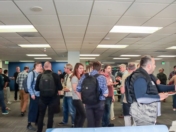 A group of people are gathered in a large, modern office space for a social event or networking mixer, with some individuals holding plates of food.