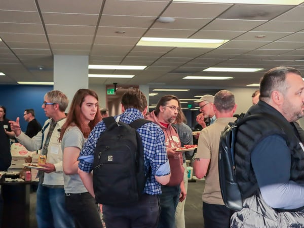 A group of people are gathered in an indoor office-like space for a social event, with several individuals holding plates of pizza and drinks while engaging in conversation.