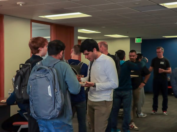 A group of people are gathered in an indoor office or common area, some wearing backpacks and holding plates of food during a social or networking event.