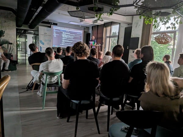 A group of people attending a technical presentation in a modern office space, with a speaker at the front and code displayed on a projector screen.