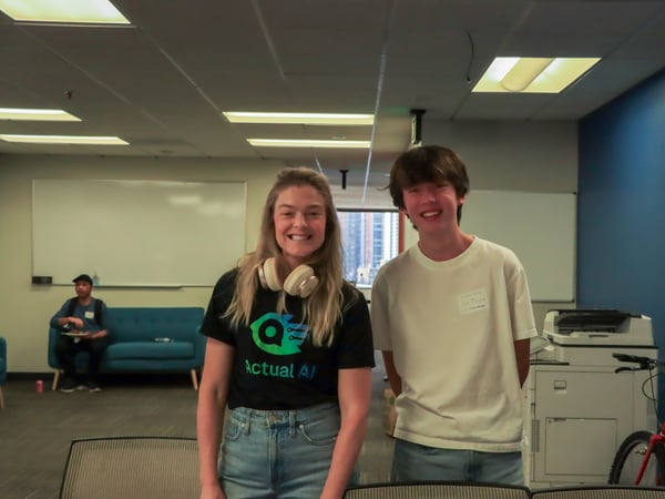 Two people, a woman and a young man, are smiling and posing for a photo in an office-like common area with a third person sitting on a couch in the background.