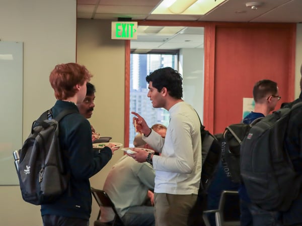 A group of people are seen socializing and eating in an indoor office or event space, with an illuminated green exit sign visible above a doorway.