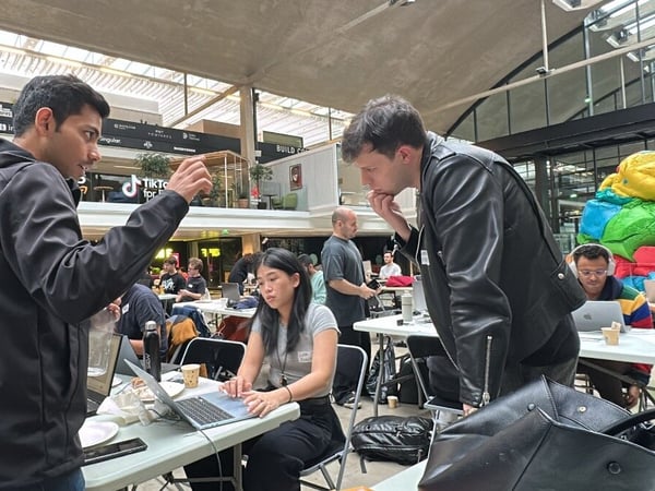A candid photograph captures two men discussing a topic while a woman works on a laptop at a table during a busy indoor tech event, likely a hackathon or workshop.