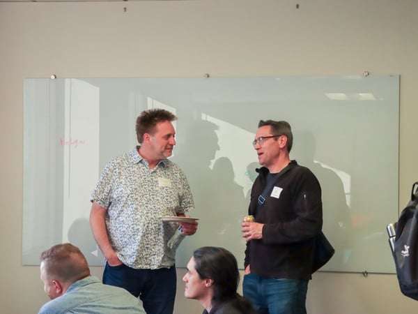 Two men are engaged in a conversation in front of a large glass whiteboard in an office or event setting.
