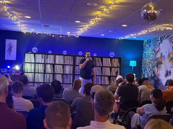 A man stands on a stage holding a microphone, performing or speaking in front of a seated audience. The background features large shelves filled with vinyl records, illuminated by blue lighting, string lights, and a disco ball.