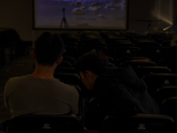 Two people sit in the foreground of a dark auditorium, looking toward a projection screen in the distance. The room is filled with rows of empty seats, and the lighting is very dim.
