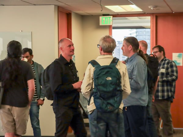 A group of men are standing and talking in an office hallway or common area near an exit.