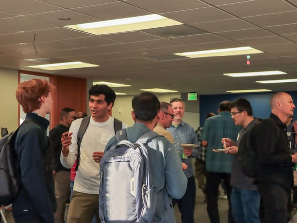 A group of people are gathered in an indoor office setting, engaged in conversation and eating during a social event.