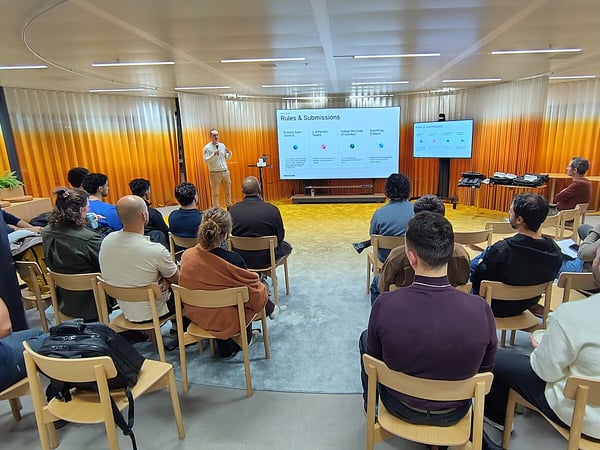 A photograph captures an indoor presentation where a speaker stands next to a large screen displaying 'Rules & Submissions' while a sizable audience is seated, facing the stage and screen.