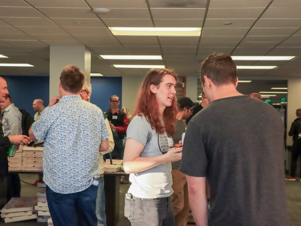 A group of people are socializing and talking in an indoor office setting with pizza boxes on a table.