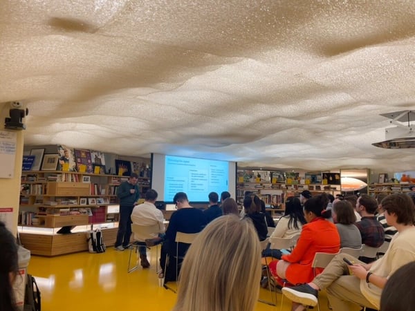A group of people attending a presentation in a modern indoor space featuring a unique, wavy textured ceiling and a bright yellow floor. A presenter stands near a projection screen displaying a slide with text and bullet points.
