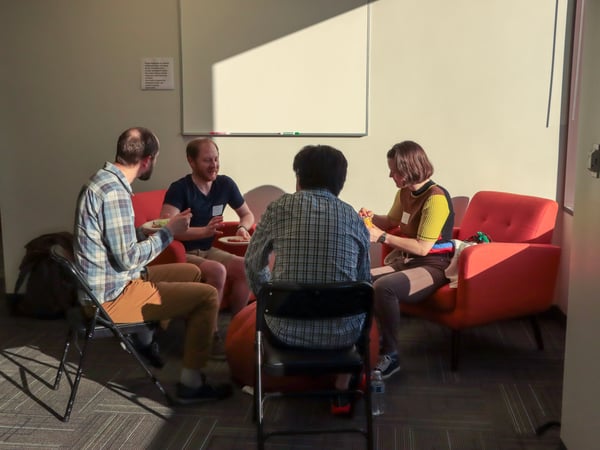 A group of people are sitting in a circle on red armchairs and folding chairs, engaged in a conversation in an indoor office or common area with a whiteboard in the background.