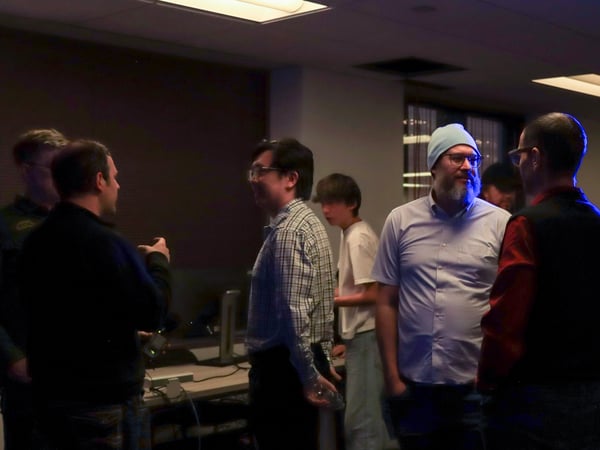 A group of men are engaged in a conversation within a dimly lit indoor setting, likely an office or event space.