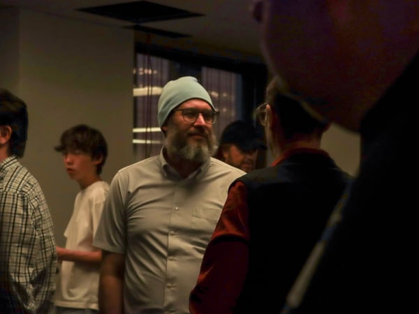 A candid indoor photograph of a group of people, featuring a man with a beard wearing a light blue beanie and glasses in the center.