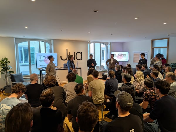 A group of people attending a tech presentation titled 'AI Tinkerers' in a modern indoor space with speakers at the front.