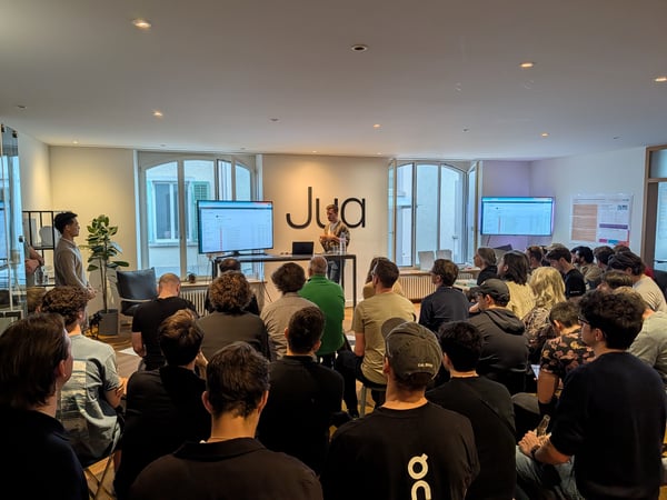 A man stands at the front of a modern office space giving a presentation to a seated audience. A large screen next to him displays a software interface, and the word 'Jua' is prominently featured on the wall behind him.