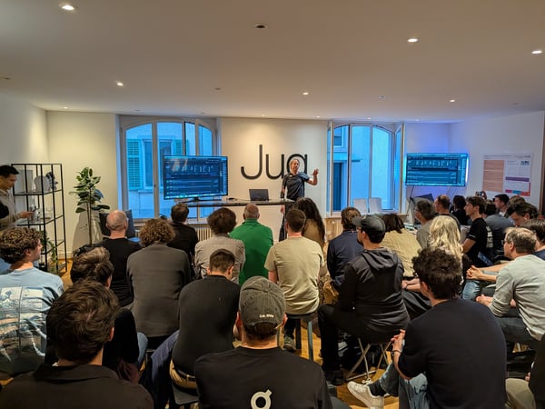 A man stands at the front of a room giving a presentation to a seated audience, with large screens and a prominent 'Jua' logo on the wall.