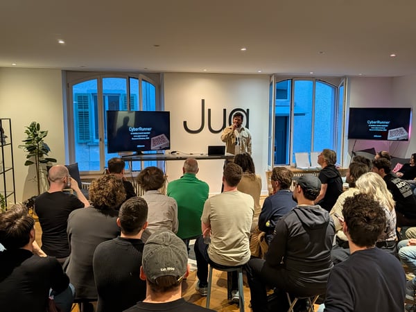 A man is giving a presentation titled 'CyberRunner' to an audience in a modern indoor space with large screens and a 'Jua' logo on the wall.