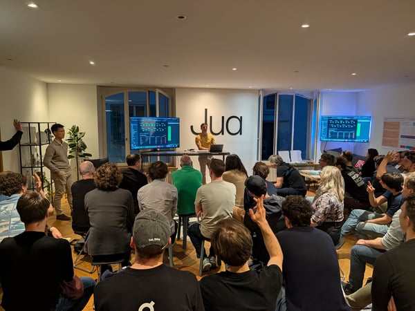 A man stands at the front of a room giving a presentation to a seated audience, with several people raising their hands to ask questions.