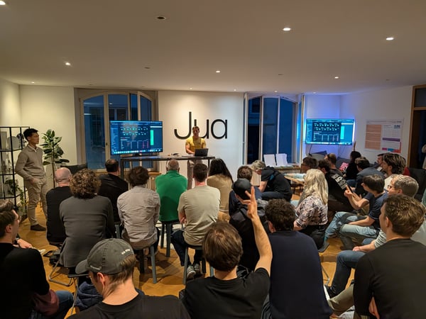 A group of people attending a tech presentation in a modern indoor space, with a presenter standing in front of a wall featuring a large 'Jua' logo.