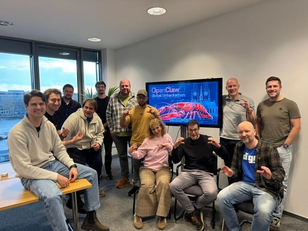 A group of twelve people posing for a photo in an office setting, many making claw-like hand gestures in front of a screen displaying 'OpenClaw Global Unhackathon'.