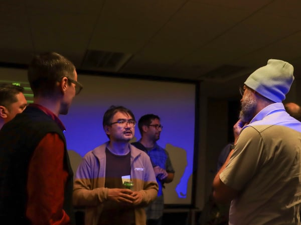 A group of people are gathered in a dimly lit room, engaged in conversation in front of a projector screen.